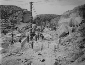 A view of the quarry face showing workers holding the tools of their trade while dressing stones. Blocks of granite are ready to be moved using the crane. Photographer: Samuel John Govier.
